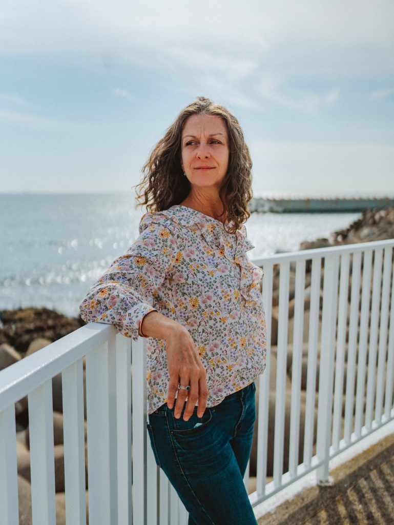A photograph of the author of the blog, looking back over her shoulder, with her arm resting on a fence in front of the ocean