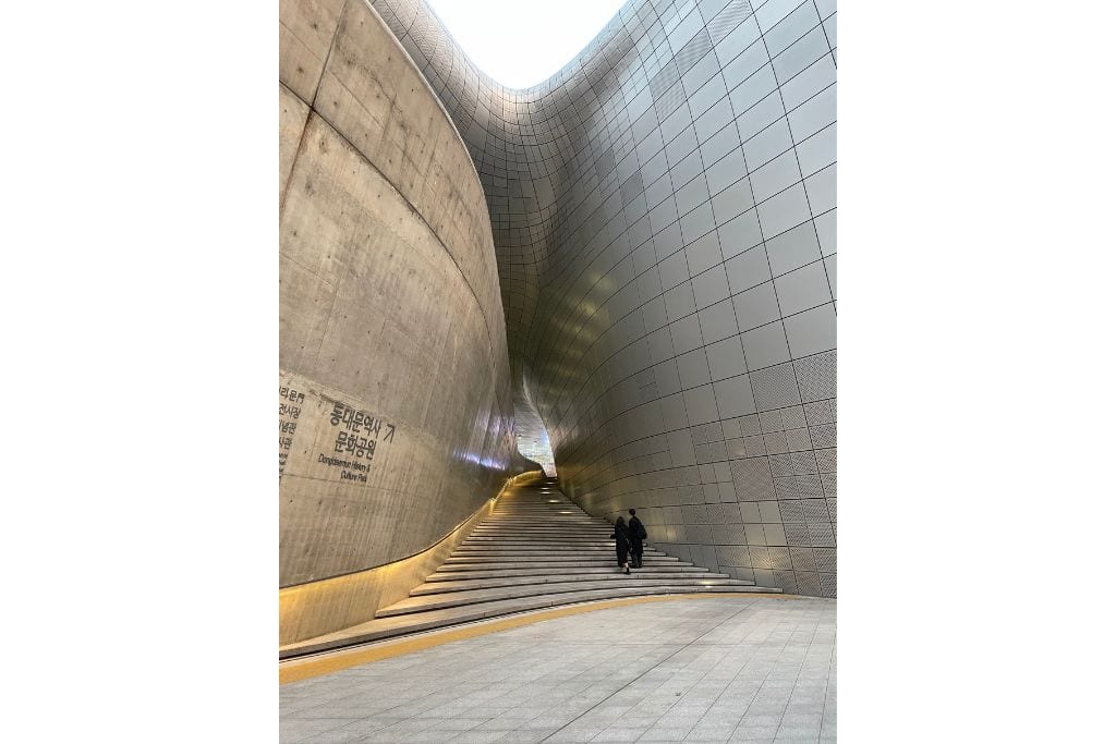 The curves of 2 buildings with stairs in between, 2 people walking up the stairs. 