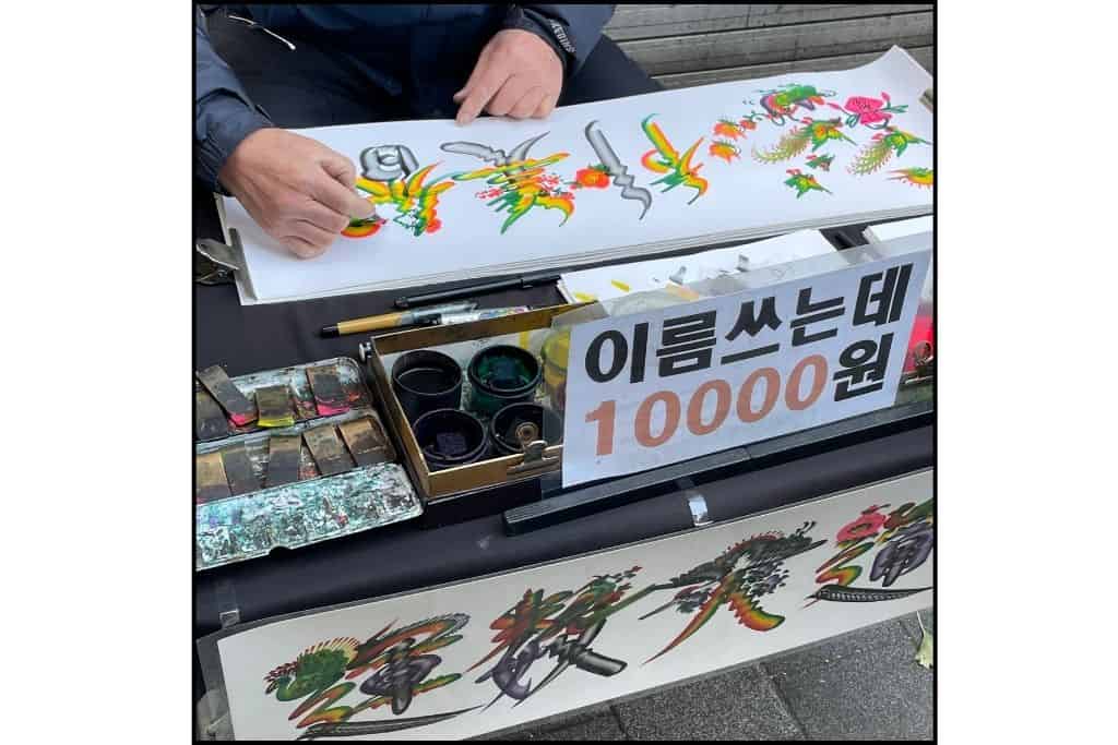 A street vendor painting a customer's name in Hangul on a piece of paper or artwork