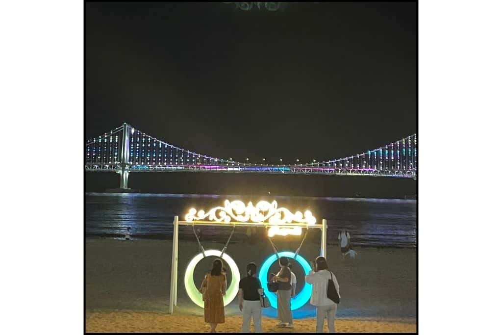 A nighttime view of Gwangan Bridge lit up with some swings that also light up at the beach