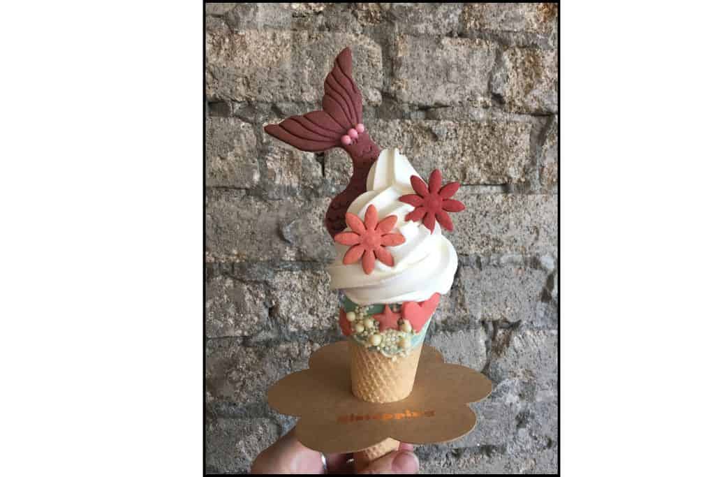 A whimsical ocean-themed ice cream cone featuring a pink mermaid tail and two pink flower-shaped cookies creates a colorful and playful design.