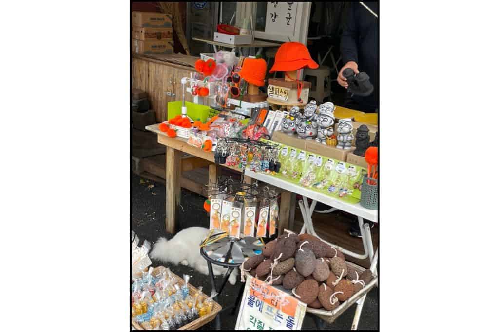 Colorful street market stall selling Jeju Island souvenirs, including Dolhareubang statues, lava stones, keychains, and (tangerine) Hallabong-themed gifts