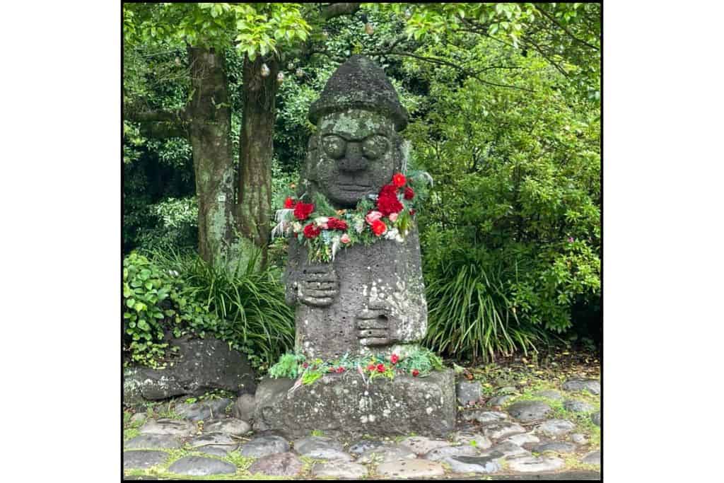 Traditional Dolhareubang stone statue decorated with red and white flower garlands, surrounded by lush greenery on Jeju Island, South Korea