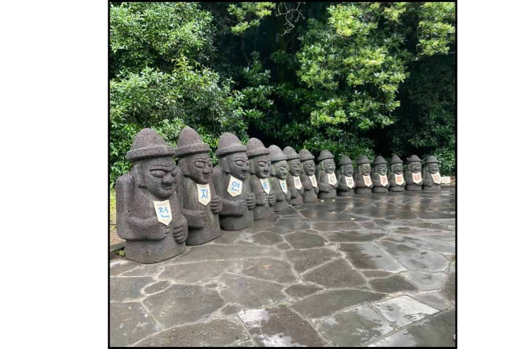 Row of Dolhareubang statues on Jeju Island, each wearing a sign with a Korean character, standing on a wet stone path surrounded by lush greenery.