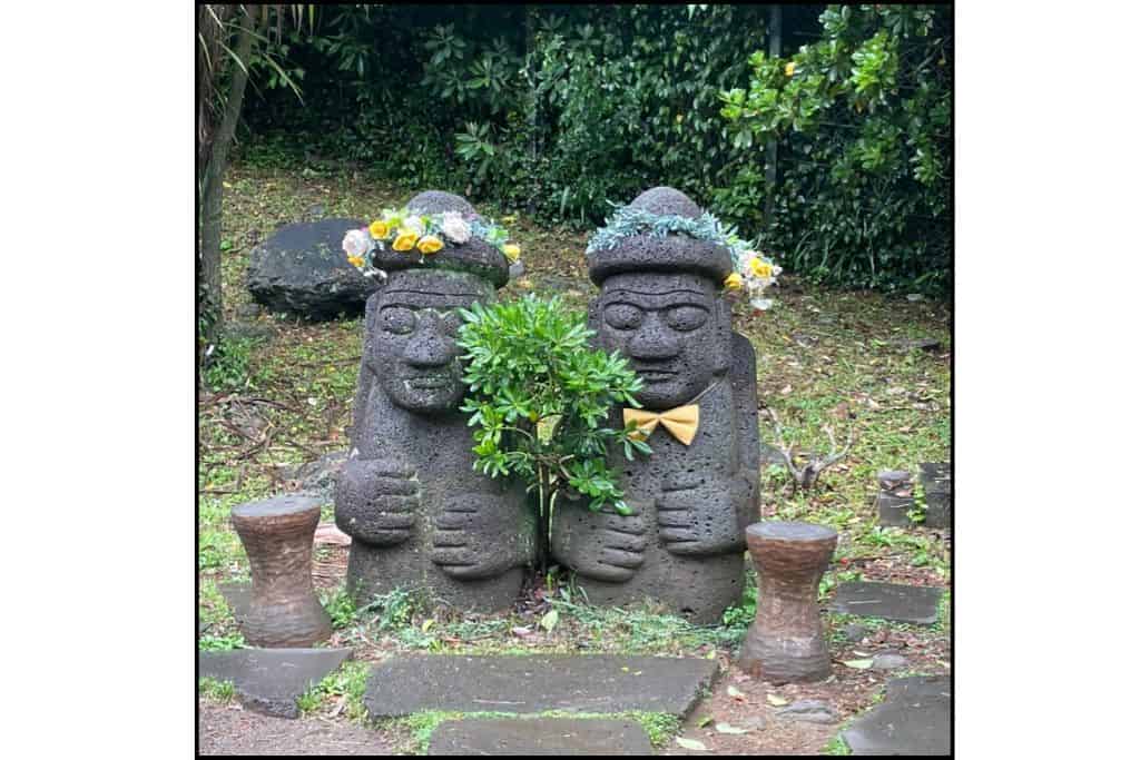 Two Dolhareubang statues on Jeju Island wearing flower crowns and a yellow bowtie, surrounded by greenery — a playful twist on the island’s traditional stone guardians.
