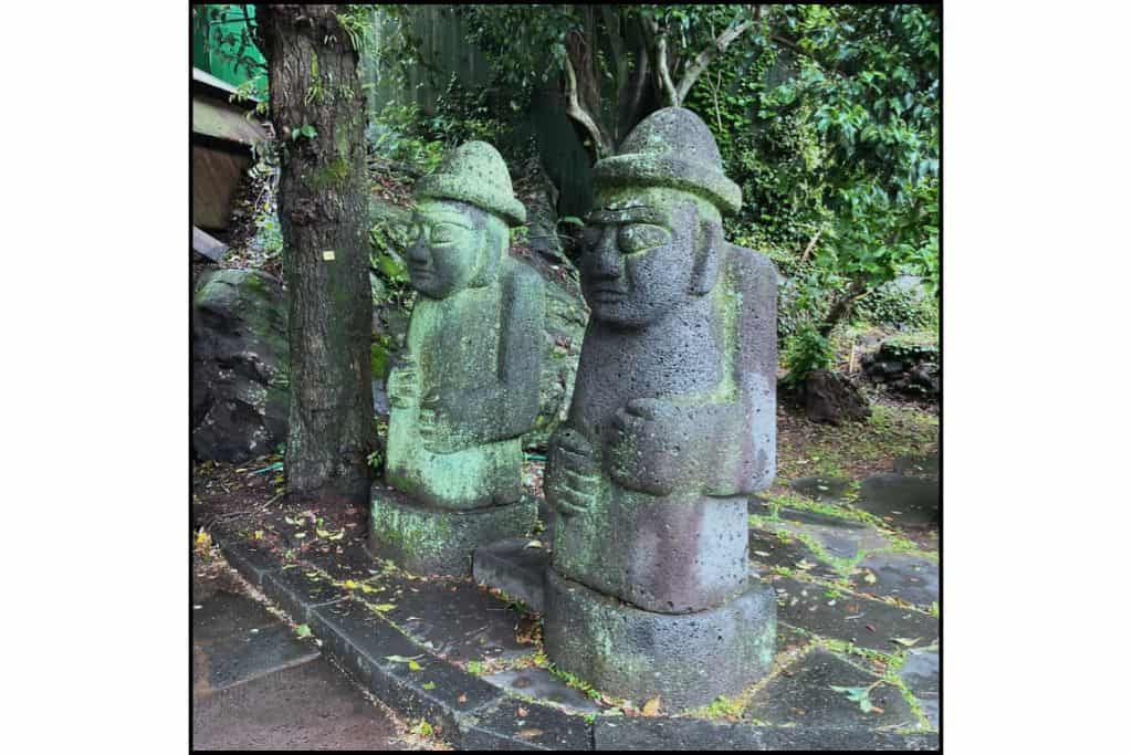 Two Dolhareubang statues on Jeju Island decorated with flower crowns and a yellow bowtie, adding a playful touch to the traditional volcanic stone guardians.