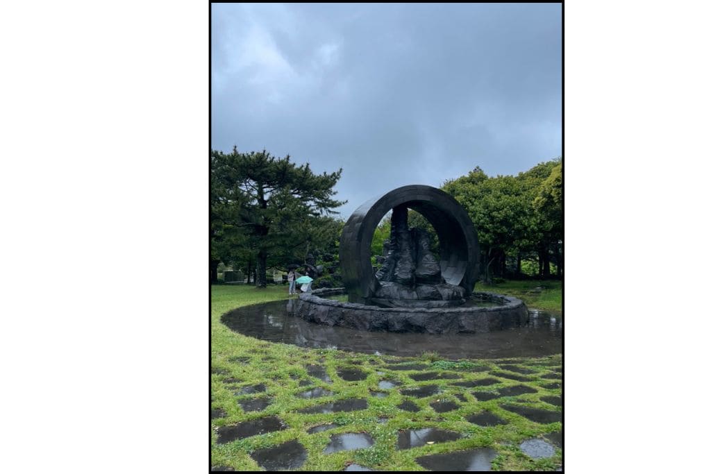 Modern stone sculpture surrounded by circular arch in a green park on Jeju Island, South Korea, with visitors holding umbrellas on a rainy day.
