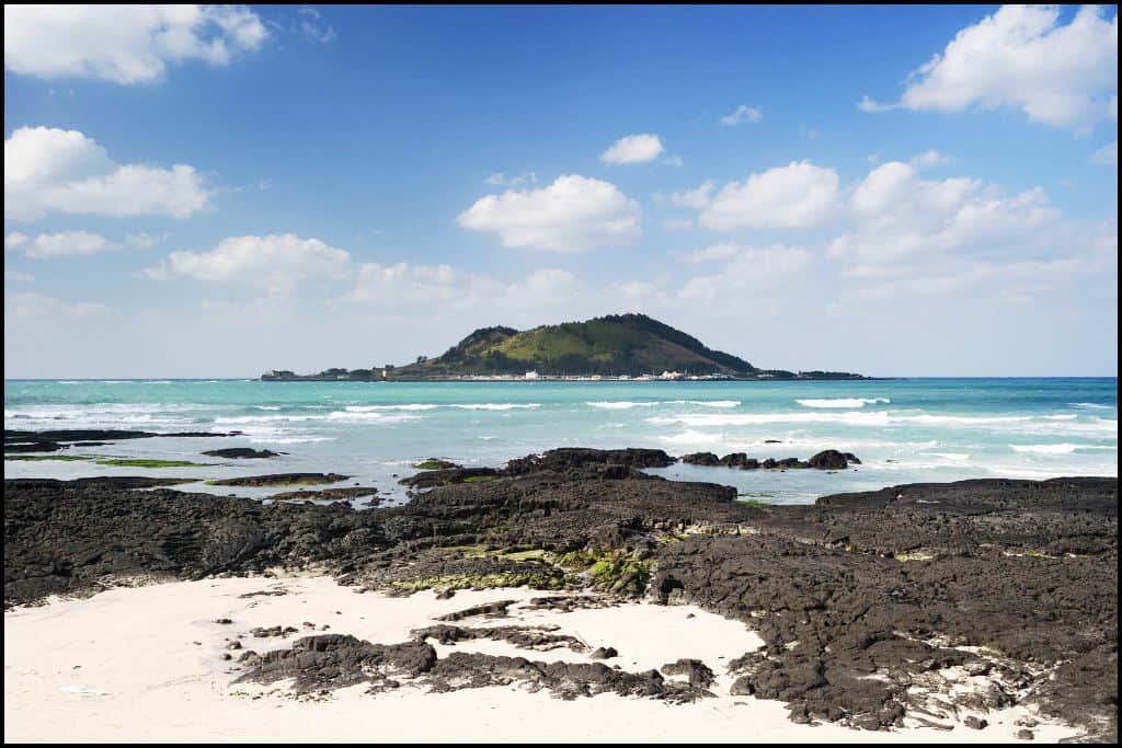 Scenic view of Hyeopjae Beach on Jeju Island, South Korea, with black volcanic rocks, soft white sand, and turquoise water facing the green, forested Biyangdo Island.