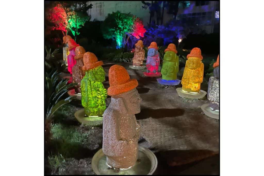 Row of illuminated Dolhareubang statues glowing in vibrant colors at night on Jeju Island, South Korea, with trees lit up in red, green, and blue in the background.