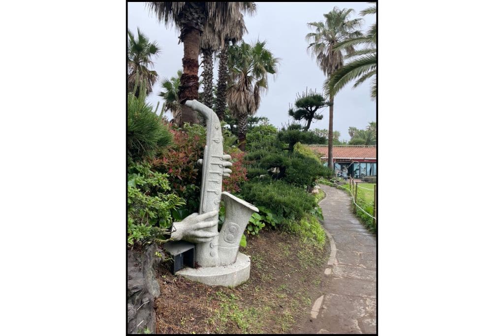 Stone sculpture of a saxophone being played by carved hands along a scenic coastal walking path lined with palm trees on Jeju Island, South Korea.