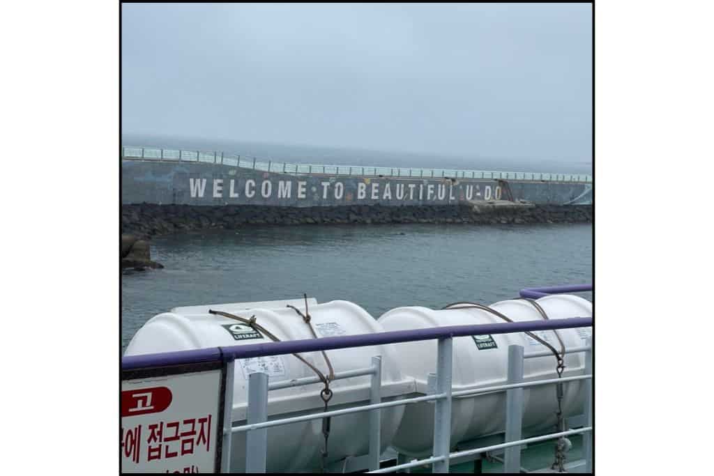 View from the ferry arriving at Udo Island, South Korea, showing the 'Welcome to Beautiful Udo' sign on the seawall under a misty sky.