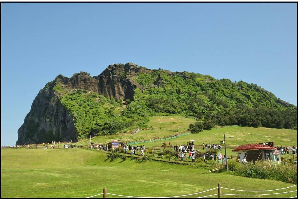 Seongsan Ilchulbong (Sunrise Peak) on a sunny day with tourists walking through grassy fields, Jeju Island