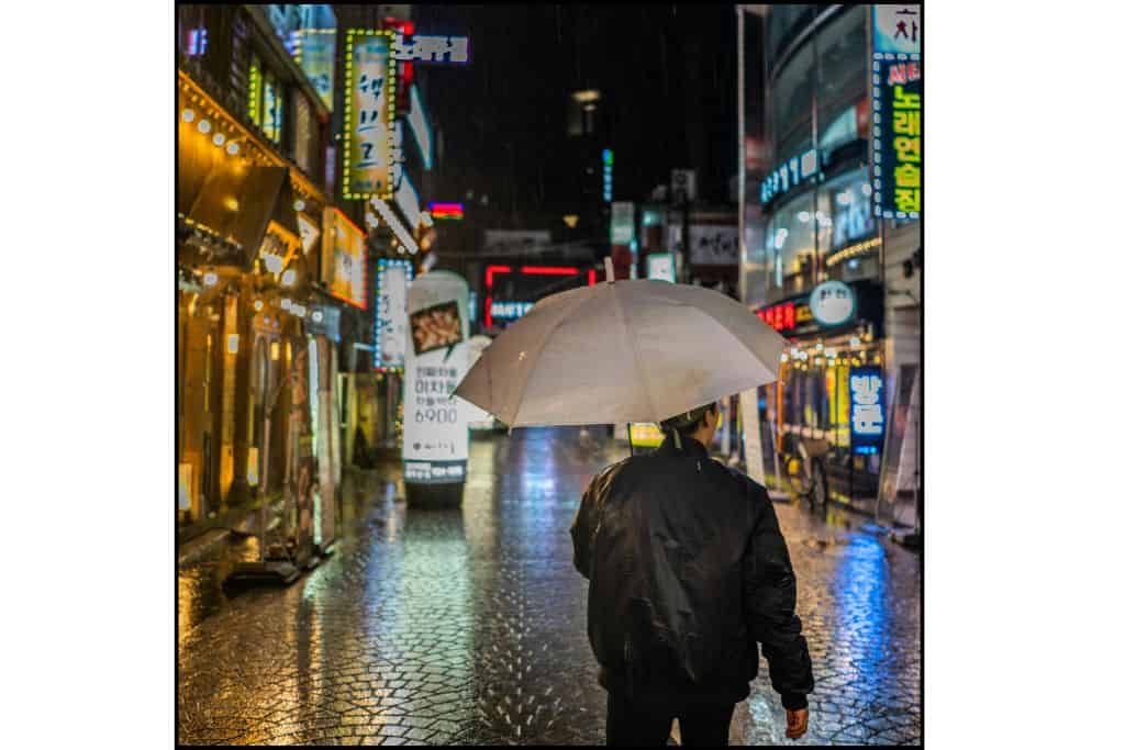 Man standing under an umbrella at night in Seoul, illuminated by streetlights."