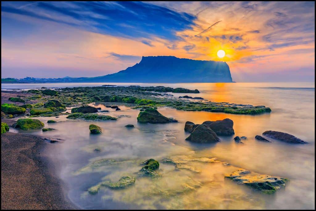 Colorful Sunrise over Seongsan Ilchulbong (Sunrise Peak) on Jeju Island, with moss-covered rocks and calm coastal waters in the foreground.
