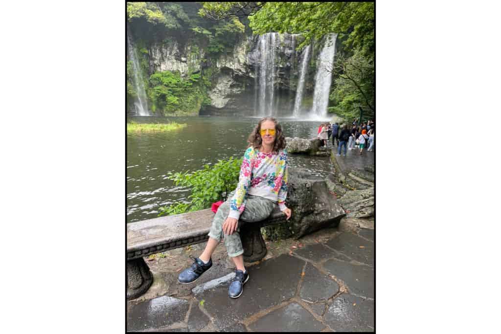 Traveler sitting on a stone bench in front of Cheonjeyeon Waterfall in Jeju Island, South Korea, wearing a colorful floral top and yellow sunglasses on a cloudy day.