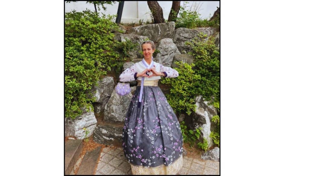 Person wearing a purple floral hanbok and making a heart hand gesture, standing in front of stone steps and greenery in Korea.