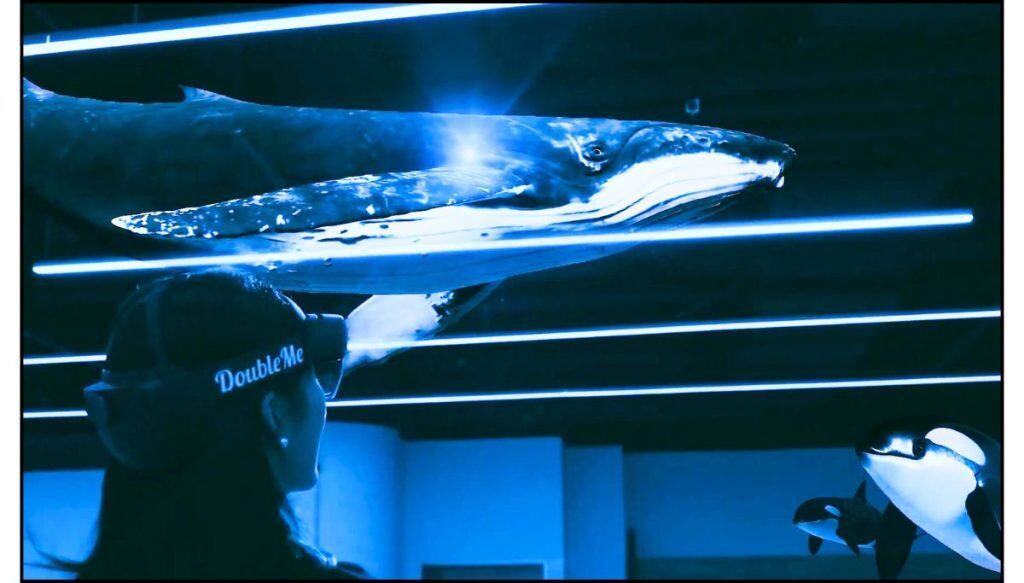 A woman wearing a VR headset at The CAVE VR Gallery in Seoul experiences a virtual underwater scene with a whale and orcas projected above.