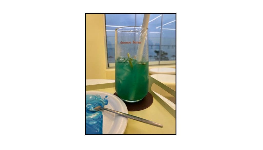 Blue iced drink with lemon slices on a yellow café table, with a plate of blue dessert and a window view in the background.