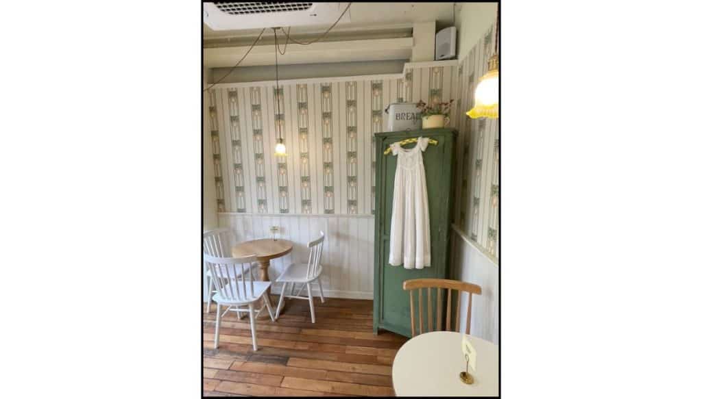 A popular cozy café's interior with wooden floors, floral wallpaper, white chairs, and a vintage green cabinet holding a white dress.