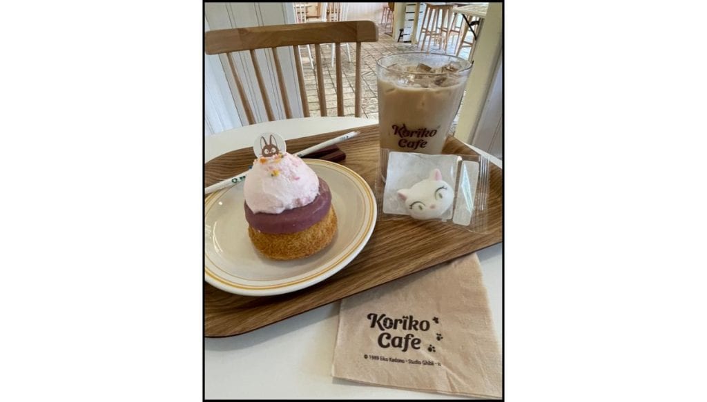 A Koriko Café dessert tray with an iced latte, pink cream puff pastry, and a cute cat-shaped treat, displayed on a wooden table.