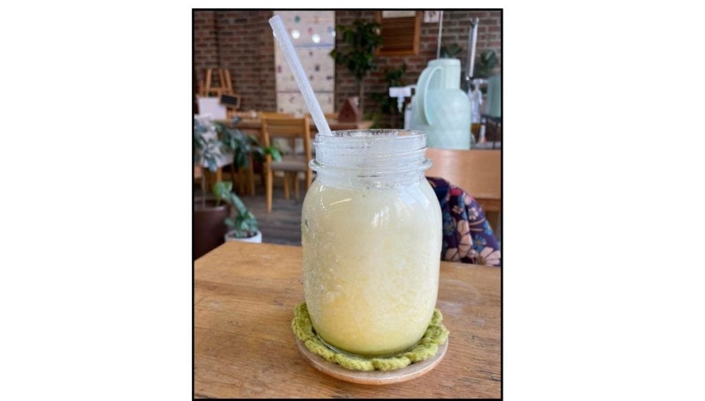 A cold drink served in a mason jar on a wooden café table with plants and cozy décor in the background.