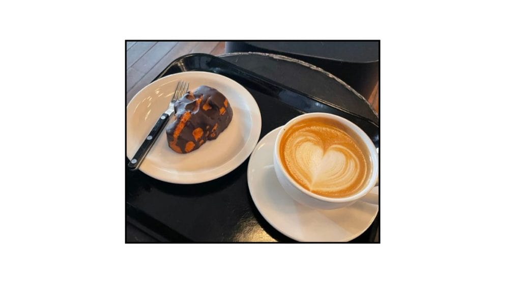 Latte with heart-shaped latte art served with a chocolate-covered pastry on a white plate and black tray.