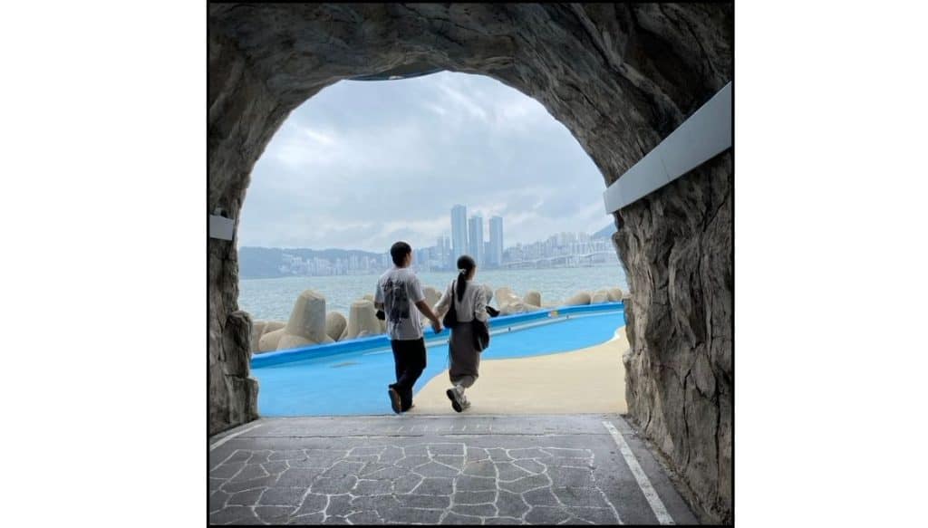 Couple walking through a rock tunnel at Huinnyeoul Culture Village with ocean and city views ahead