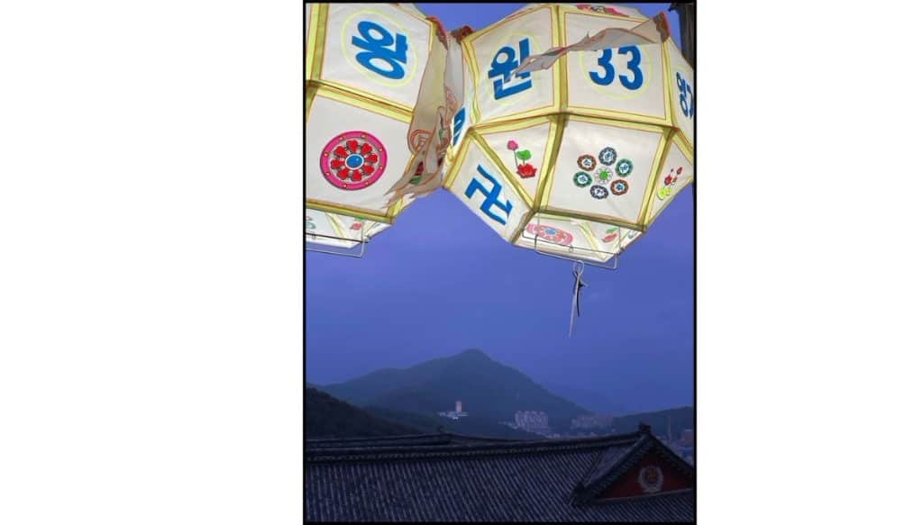 Hanging lotus lanterns with Korean characters above Samgwangsa Temple rooftops, with mountains visible in the distance at dusk.