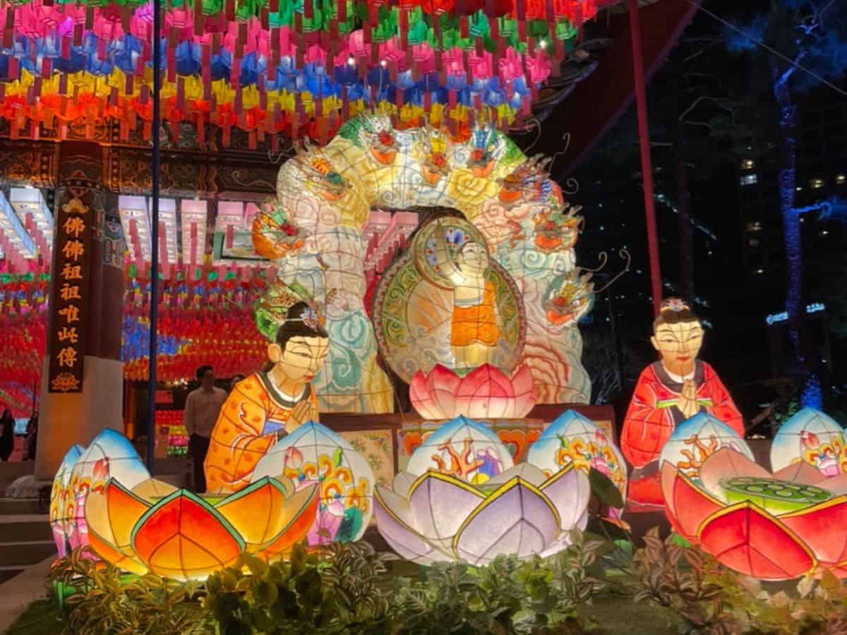 At night, large illuminated lantern sculptures depict seated Buddhist figures and lotus flowers in red, pink, and white, arranged in front of a temple pavilion. Hundreds of small, colorful paper lanterns hang overhead, filling the scene with bright light and color.