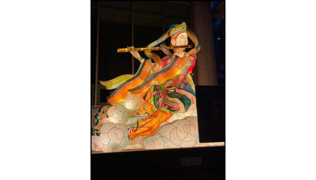 Illuminated lantern figure depicting a robed Buddhist celestial musician playing a flute, shown floating above stylized clouds, with flowing garments in shades of orange, green, and gold glowing against a dark nighttime background.