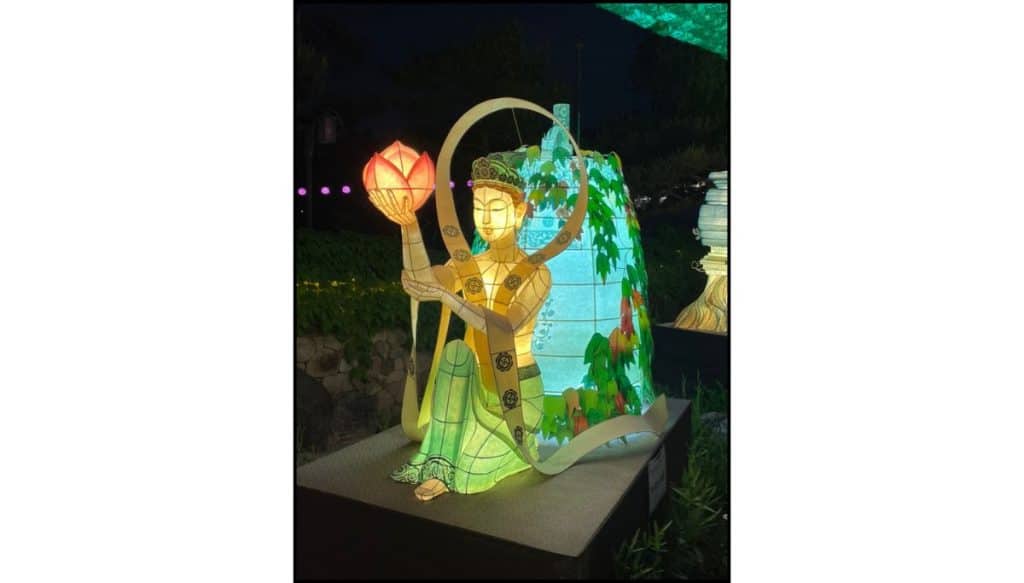 Illuminated lantern sculpture of a seated Buddhist figure holding a glowing lotus flower at night, with green and gold robes, a halo-like frame, and leafy decorative panels behind it in a garden setting at night