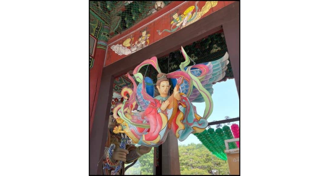 A colorful hanging lantern sculpture depicts a winged figure with flowing ribbons, suspended beneath a painted wooden temple gate during daylight. Rows of green lanterns and trees are visible in the background.
