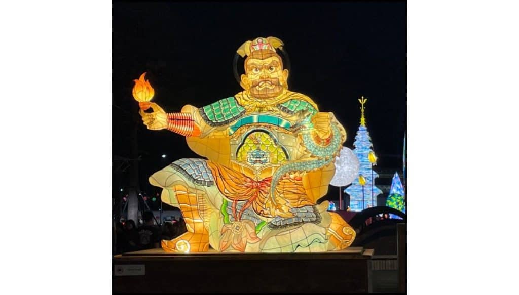 At night, a large illuminated lantern sculpture depicts a seated armored figure holding a flame in one hand, glowing in shades of yellow, orange, and green. Other colorful lantern displays are visible in the background against the dark sky.