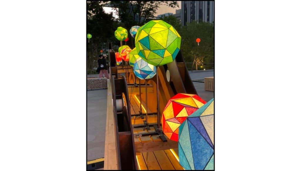 Colorful geometric lantern spheres in green, blue, yellow, and red glow along a wooden walkway at dusk, with people strolling nearby and city buildings and trees in the background.