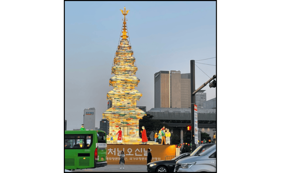 Tall illuminated lotus lantern tower made of stacked book-shaped panels stands in Gwanghwamun Square at dusk, with small colorful human figures at the base and city traffic and high-rise buildings surrounding it at dusk.