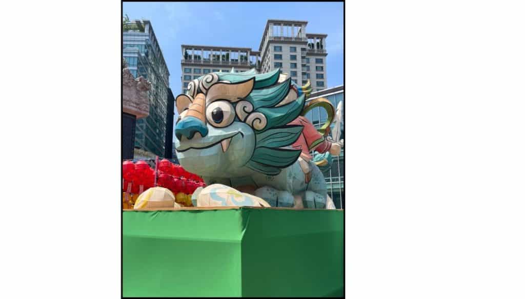 Large colorful lantern sculpture shaped like a playful guardian creature with teal and gold details, displayed outdoors on a green platform, with red lanterns and modern city buildings in the background under a clear blue sky.