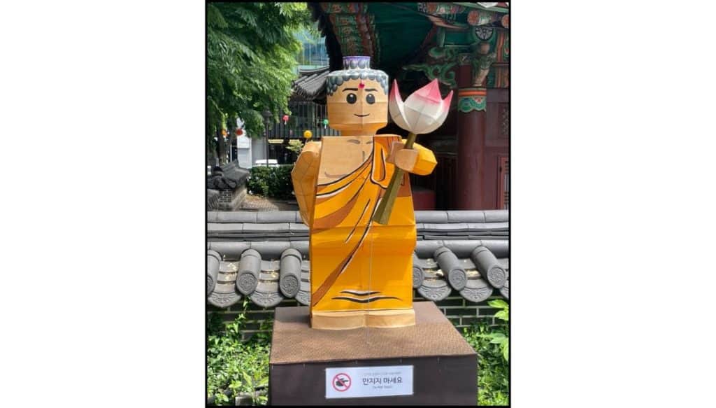 Lego-style lantern sculpture of a smiling Buddha holding a pink lotus flower, wearing an orange robe, displayed outdoors in front of a traditional Korean temple with tiled roofs and green trees in the background.