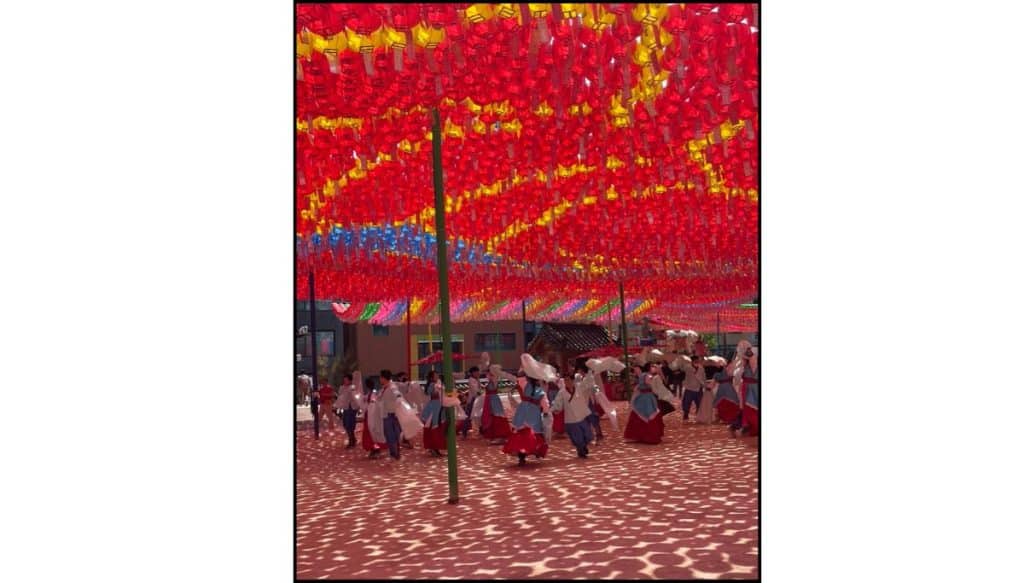 Group of performers in traditional Korean hanbok dance together at Jogyesa Temple beneath a canopy of red, yellow, and blue lotus lanterns, with patterned sunlight casting shadows across the temple courtyard.