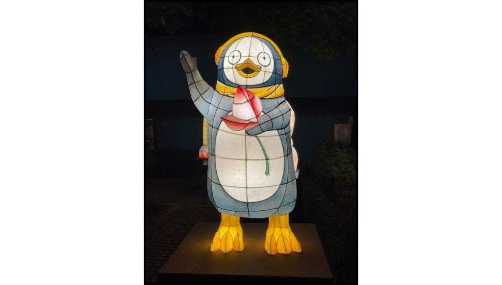 Illuminated lantern sculpture of a cartoon-style penguin wearing headphones, waving one flipper and holding a pink lotus flower, glowing at night against a dark outdoor background.