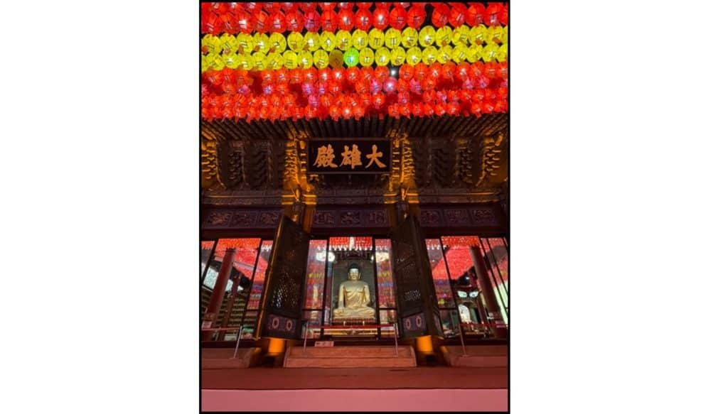 Illuminated Buddhist temple hall at night with rows of red and yellow lanterns hanging from the ceiling, glowing above a seated golden Buddha statue framed by open wooden doors and ornate carved pillars.