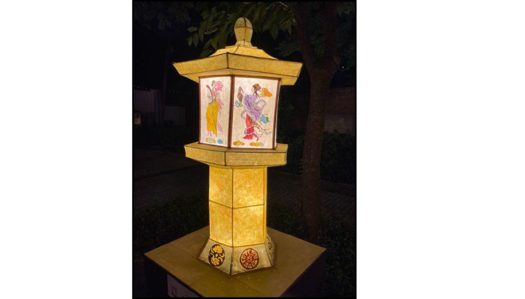 Illuminated lantern sculpture resembling a stone pagoda, glowing warm yellow at night, with painted panels showing Buddhist celestial figures dancing against a dark garden background.