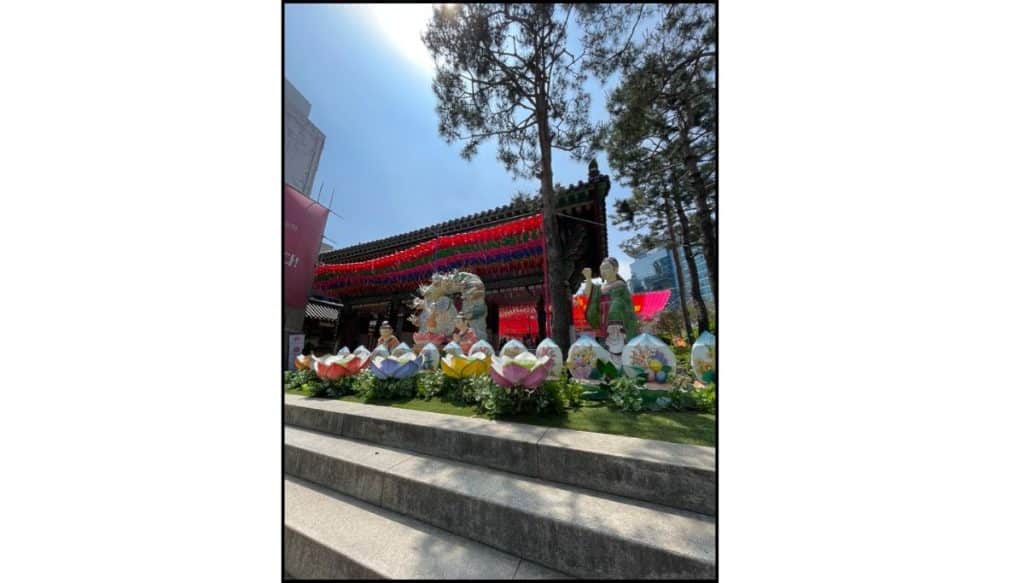 Daytime view of lantern sculptures and lotus-shaped lantern flowers arranged on steps outside Jogyesa Temple, with colorful lanterns hanging across the temple entrance and tall trees framing the scene.