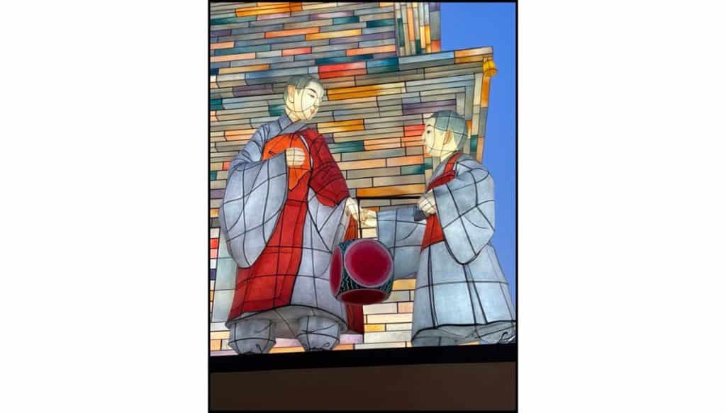 Illuminated lantern panel showing two Buddhist monks in gray and red robes facing each other and sharing a lantern, set against a mosaic-style background of glowing rectangular panels at dusk.