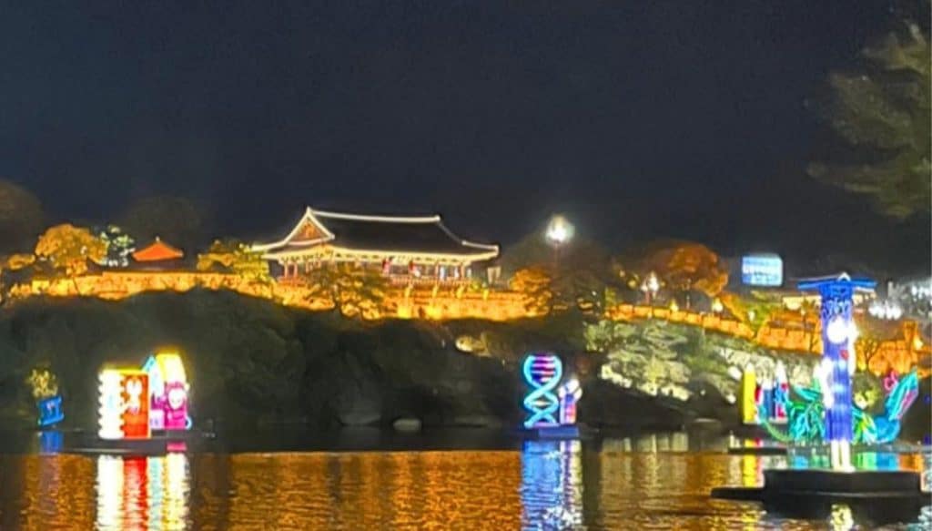 Chokseokru Pavilion lit up at night above the Jinjuseong Fortress walls, with colorful lanterns floating on the Nam River during the Jinju Namgang Yudeung Festival