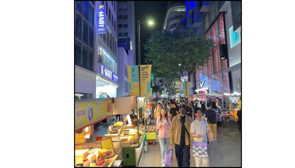 Crowded Myeongdong night street with food stalls, shoppers, and brightly lit storefronts.