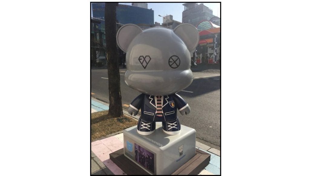 K-Star Road GangnamDol bear statue themed for K POP band EXO, displayed on a sidewalk pedestal near the street.
