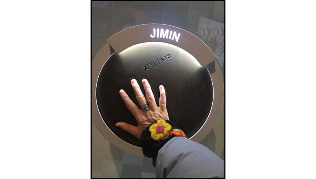 Hand touching Jimin BTS handprint display plaque, with signature visible and a colorful flower cuff on the wrist.