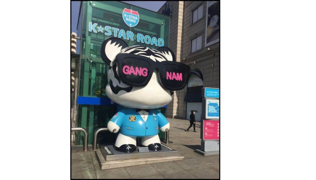 Large K-Star Road bear statue wearing sunglasses with Gang Nam text, standing beside a street entrance sign.