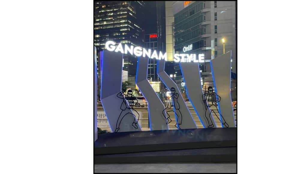 Gangnam Style stage in Seoul with illuminated sign, dancing figure silhouettes, and city buildings at night near Gangnam Station.