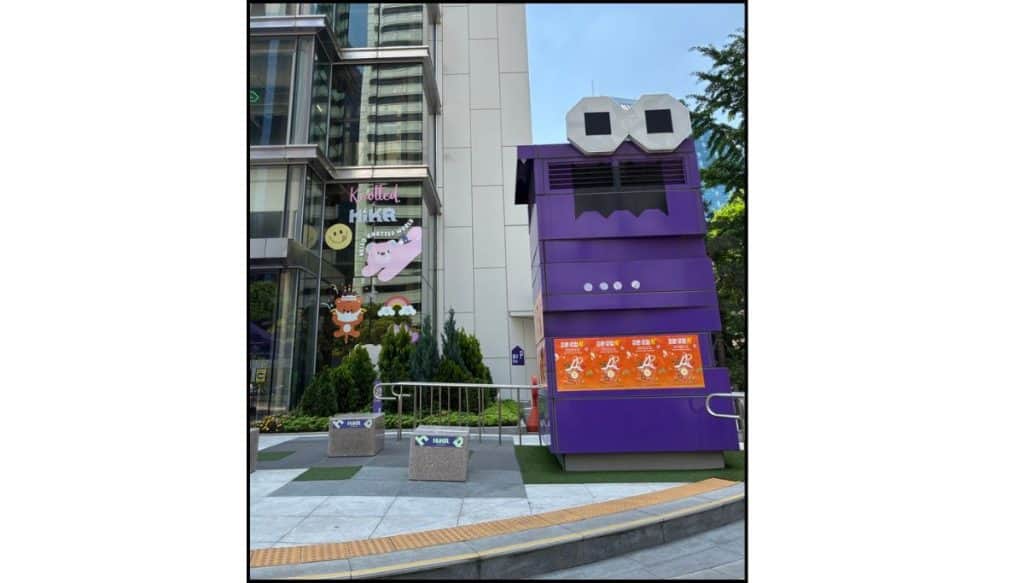 Purple HiKR character-shaped structure outside the Korea Tourism Organization building, beside glass windows with cartoon decals.